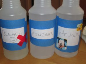 Close up of 3 spray bottles to see labels. Labels from left: Bleach with frowny face and red x, Vinegar, Helpers with mickey mouse stickers