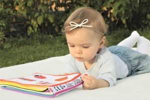 picture: baby girl laying on blanket outside looking at toy book