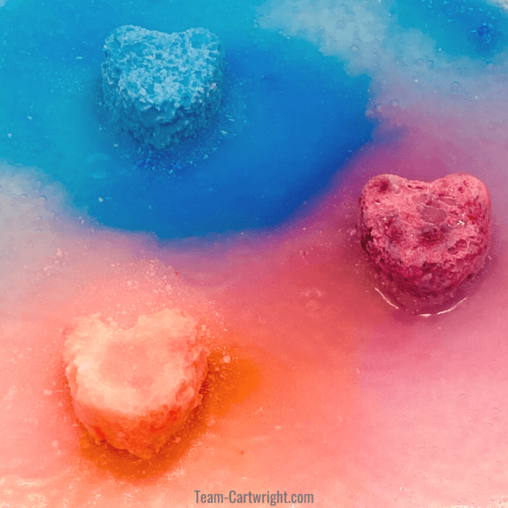 picture of a blue heart, purple heart, and pink heart fizzing as a result of the fizzy heart science experiment. Colored liquid surrounds the hearts